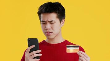 Asian young man in a red sweater holds a credit card in one hand and a smartphone in the other, displaying a thoughtful expression while engaged in an online transaction. video