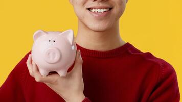 Asian young man smiles while adding a coin to his pink piggy bank, showcasing the importance of saving money. The vivid yellow backdrop highlights the cheerful atmosphere of financial responsibility. video
