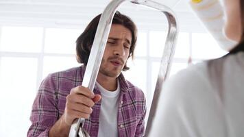A man stands on a ladder, pretending to sing joyfully as he gets ready to paint a wall. Another person is holding a paint roller, adding to the fun atmosphere of home improvement. video
