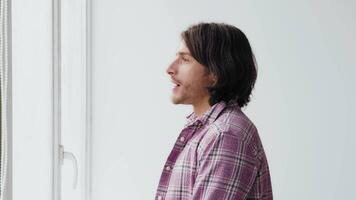 A young man with long dark hair looks thoughtfully out a large window while standing in a bright, minimalistic room. Natural light enhances his introspective moment. video