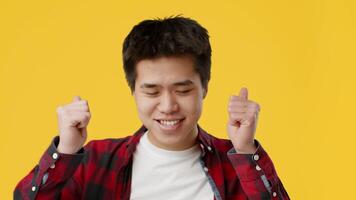 A young Asian guy in a checkered shirt smiles confidently while pointing towards the right. The vibrant yellow backdrop enhances the cheerful atmosphere of the moment. video