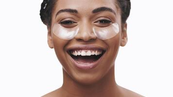 African American woman enjoys her self-care routine, smiling while applying under-eye patches. Her joyful expression reflects a moment of relaxation and self-pampering. video