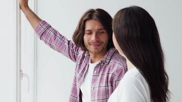 A young man and woman engage in a friendly conversation near a window, illuminated by soft, natural light. Their expressions convey warmth and connection in a relaxed atmosphere. video