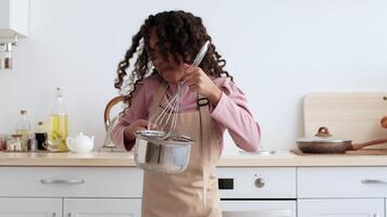 Black girl wearing an apron enthusiastically mixes ingredients in a bowl with a whisk, surrounded by kitchenware in a cozy, well-lit kitchen setting, showcasing her baking skills. video