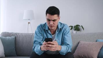 A young man is seated on a cozy couch in a light-filled room, intently looking at his smartphone. The atmosphere is calm and inviting, with soft decor elements around him. video