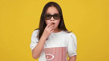 Asian child is munching on popcorn, wearing 3D glasses, with a bright yellow backdrop adding a playful touch. video