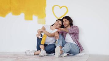 A couple relaxes against a partially painted wall, sharing a lighthearted moment while applying bright yellow paint. They seem joyful and content, reflecting a creative home project. video