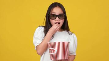 Asian girl is surprised while reaching into a bucket of popcorn, wearing 3D glasses, with a bright yellow backdrop. video
