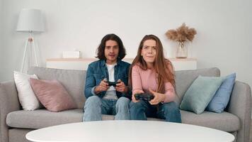 A young couple sits closely together on a soft sofa, focused on their gaming controllers video