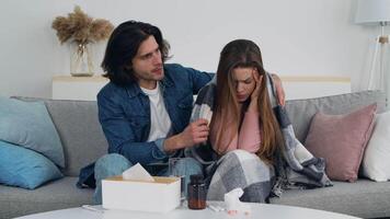 A man gently drapes a blanket over a woman sitting on the couch, who appears unwell. Tissues and medicine are nearby, indicating a cozy yet difficult day at home. video