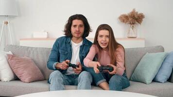 A young couple sits on a comfortable couch, intensely focused on their game controllers. Their expressions reflect excitement and surprise as they play together in a bright, inviting room. video