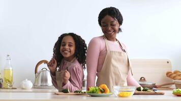 Black mother and daughter, are in a kitchen preparing food. The mother is smiling while looking over her shoulder at her daughter, who is laughing while cutting up a piece of produce. video