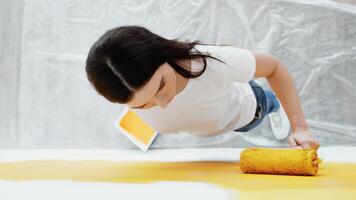 A woman is applying vibrant yellow paint to a wall while standing on a covered floor. She holds a paint roller in her hand and focuses on creating an even coat, enhancing the interior space. video