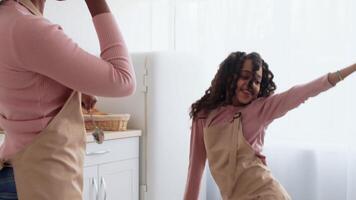 African American young girl with curly hair dances joyfully while cooking with her mother in a well-lit kitchen. They wear matching aprons and share a fun, engaging culinary experience. video