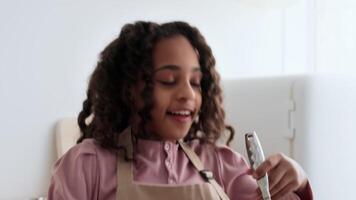 A cheerful black girl with curly hair is joyfully mixing ingredients in a bright kitchen. She wears an apron and has a big smile, enjoying her time while baking. video