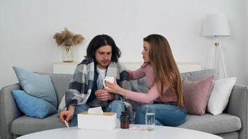 A woman comforts a sick man sitting on a couch in a modern living room, offering him a cup. They share a warm moment, surrounded by a cozy atmosphere and soft decor. video