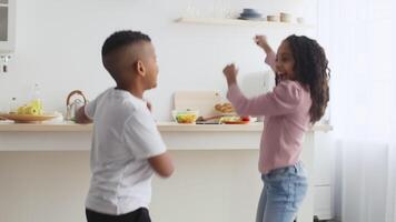 Two black children are happily dancing in a sunlit kitchen, surrounded by snacks and fruit. Their energy and excitement create a lively atmosphere in the cozy space. video