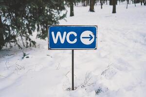 Sign indicating toilet location on a snowy path in a wooded area during winter photo