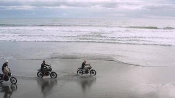 Three handsome hipster men riding modern custom motorcycle cafe racer on the black sand beach near the ocean. Adventure travel concept. Surfing spot with ocean waves. Aerial shot. 4k video