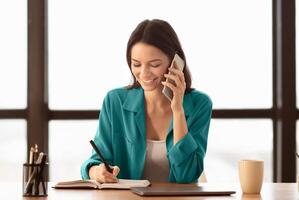 Smiling business woman talking on phone and taking notes, sitting at her working place in office, copyspace photo