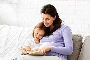 Friendly intelligent family pregnant mother and little daughter reading book on couch at home, empty space photo