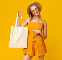 Mockup For Design. Cheerful Summer Girl Holding White Eco Tote Bag And Pointing At It, Posing Over Yellow Background With Empty Space photo