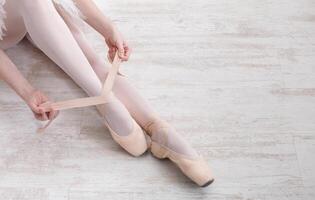 Beautiful legs of young ballerina who puts on pointe shoes at white wooden floor background, top view from above with copy space. Ballet practice. Beautiful slim graceful feet of ballet dancer. photo