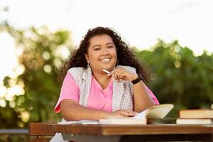 A young student sits at a wooden table surrounded by greenery, smiling while holding a pen and studying from an open book under the warm sunlight. photo
