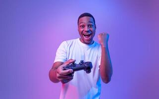 Gaming Concept. Euphoric African American Guy With Joystick Celebrating Game Win, Cheerful Black Man Raising Fist And Exclaiming With Excitement, Standing In Neon Light Over Purple Background photo