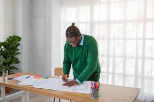 A black man is focused on sketching designs with colored markers on a large sheet of paper at a well-lit wooden desk. The workspace features natural light and greenery. photo