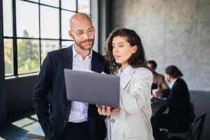 Two colleagues engage in a collaborative discussion while reviewing a laptop. The backdrop features a contemporary office with a large window and a team in the background. photo