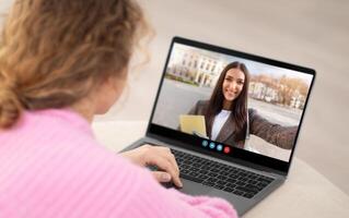 A woman interacts with a friend on a call, smiling and holding a yellow envelope. She sits at a table, with a laptop displaying her friend's cheerful face and a blurred outdoor background. photo