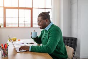In a well-lit office, a black man in a green sweater sips coffee while focusing on his laptop. The setting features a clean desk with stationery and fresh fruit, creating an inviting atmosphere. photo