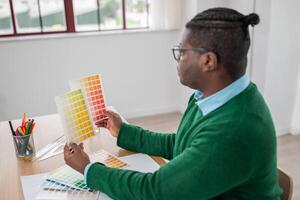A black man sits at a desk in a bright workspace, carefully comparing color swatches. Pencils and tools are organized nearby, showing his focus on a design project. photo
