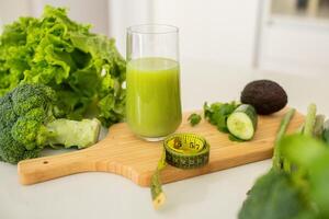 A vibrant green smoothie sits in a glass on a cutting board surrounded by fresh broccoli, avocado, lettuce, cucumber, and measuring tape, highlighting a nutritious lifestyle choice. photo