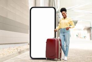 A cheerful black lady traveler in a yellow shirt and jeans stands beside a large blank screen, pulling a red suitcase. The setting suggests a modern travel hub, showcasing preparation for a journey. photo