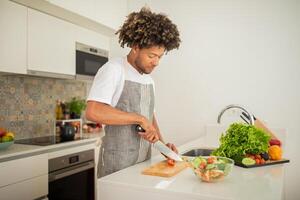 A black man is chopping tomatoes and preparing a salad in a stylish kitchen filled with fresh vegetables. The bright space highlights a relaxing cooking atmosphere during the day. photo