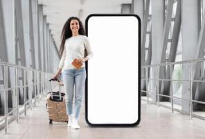 A young woman is walking confidently in a modern corridor, pulling her suitcase behind her while holding a travel document. A large blank screen stands beside her, creating a striking backdrop. photo