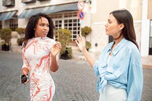 Two women are engaged in a lively conversation on a cobblestone street, surrounded by charming buildings and greenery. Their expressions convey interest and excitement. photo