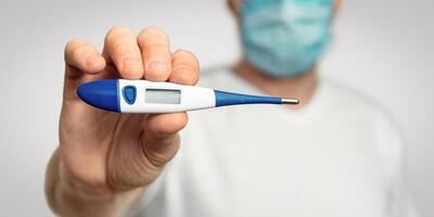 Coronavirus Disease Symptom. Sick Man Having Fever Showing Thermometer Over Gray Background, Wearing Protective Mask. Selective Focus, Shallow Depth photo