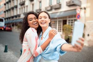 Two smiling friends pose together while taking a selfie on a vibrant urban street during daylight. The lively atmosphere enhances their friendship and happiness. photo
