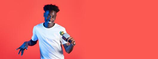 A joyful young black man confidently holds a microphone, striking a playful pose against a bright red background. His expression and stance convey fun and creativity in a lighthearted atmosphere. photo
