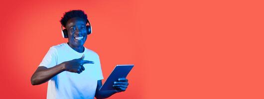 A young black man smiles while listening to music on headphones and pointing at a tablet in a vibrant, colorful studio setting. His joyful expression suggests he is enjoying himself immensely. photo