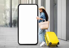 A young woman stands in a sleek urban plaza, featuring a large blank screen. She carries a yellow suitcase and a woven basket, enjoying her coffee while embracing her travel adventure. photo