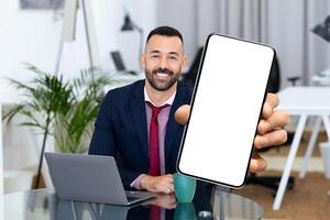 A man in formal attire sits at an office desk, smiling and holding a smartphone in front of him. His laptop and a cup are nearby, creating a productive atmosphere. photo