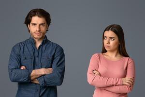Young woman looking with sorrow at her offended husband after their argue, couple standing with folded arms over grey background, empty space photo