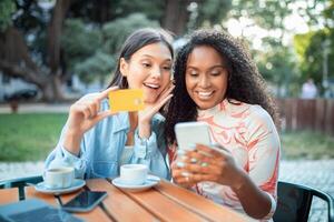 Two young women are seated at a table outside, happily engaging with their smartphones while sipping coffee. They are in a park setting filled with trees, creating a lively atmosphere. photo