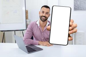 A man with a friendly smile holds up a blank smartphone in a bright office. A laptop is placed beside him, creating a workspace ambiance suitable for digital activities. photo