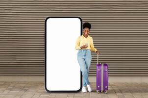A cheerful young black woman stands next to a large blank smartphone display, holding a mobile device. She has a purple suitcase and is dressed casually, enjoying the outdoor atmosphere. photo