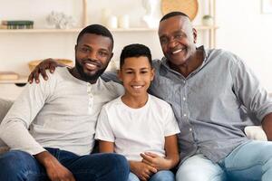 Portrait of smiling millennial black dad, preteen son and grandfather posing to camera at home, sitting on couch in living room, smiling photo